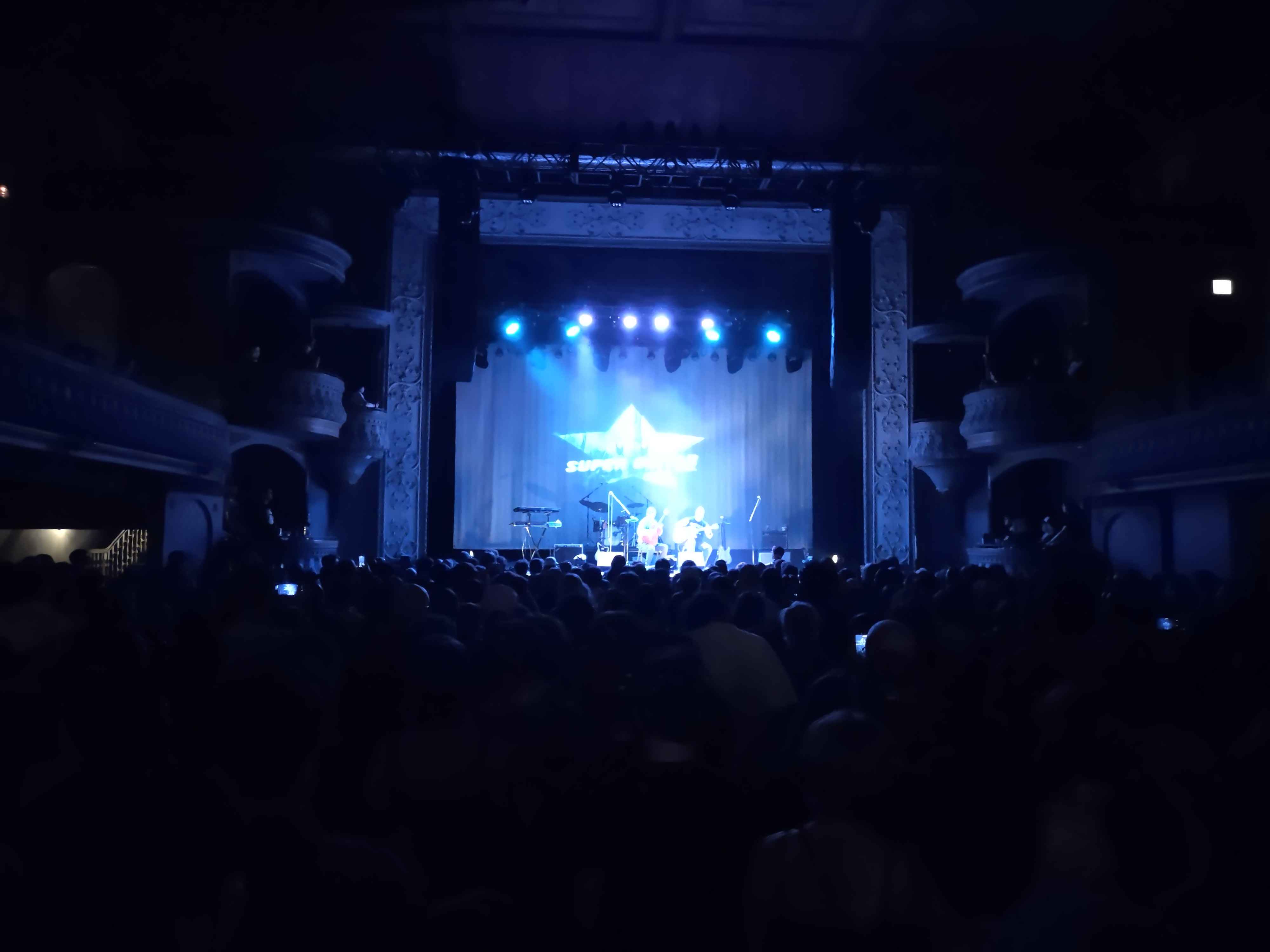 a photo taken of a stage, two guitar players standing in front of a crowd under blue and purple lights.