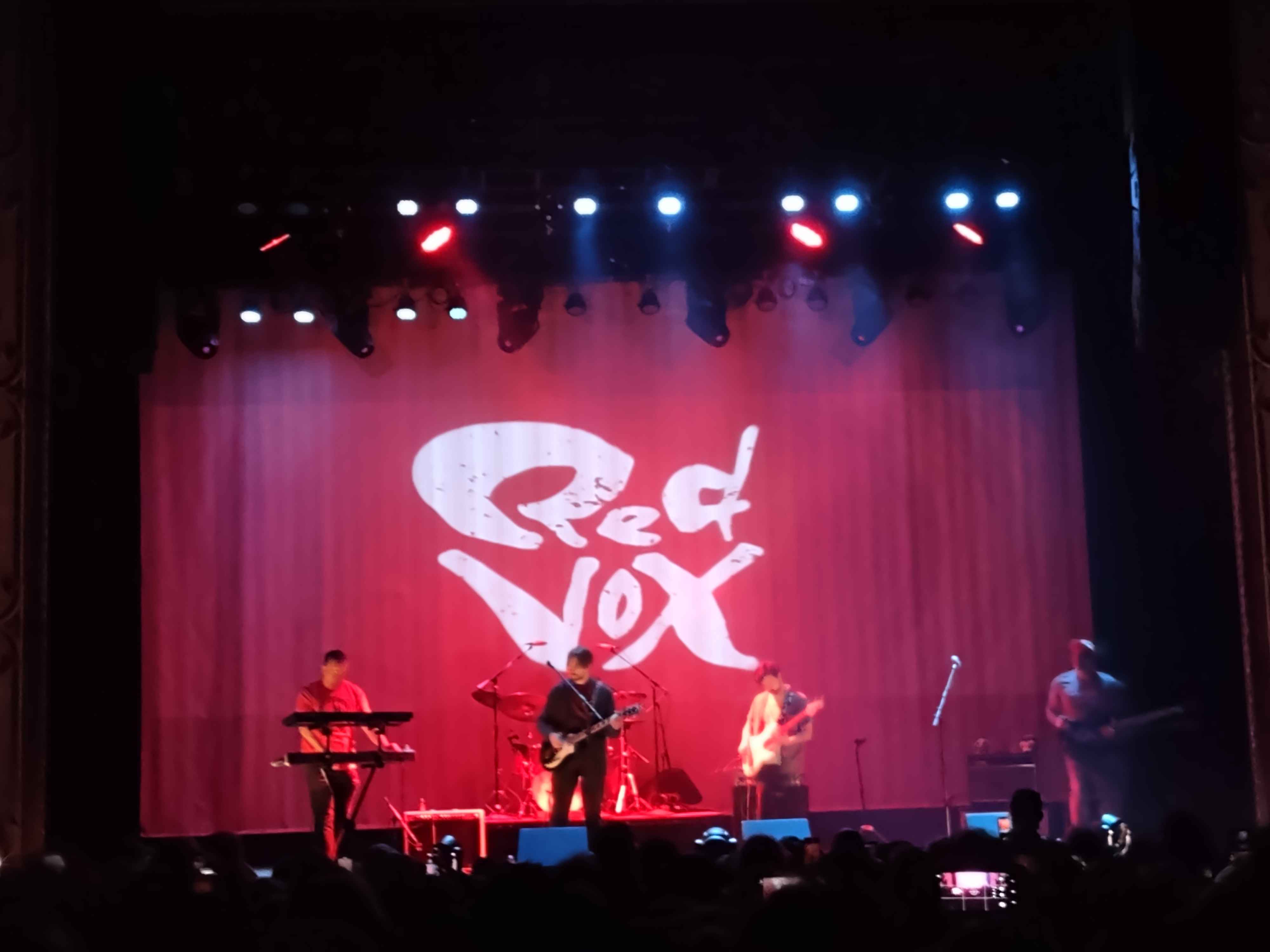 a photo of a six piece band with the drummer obscured playing in front of a crowd under red and blue lights.