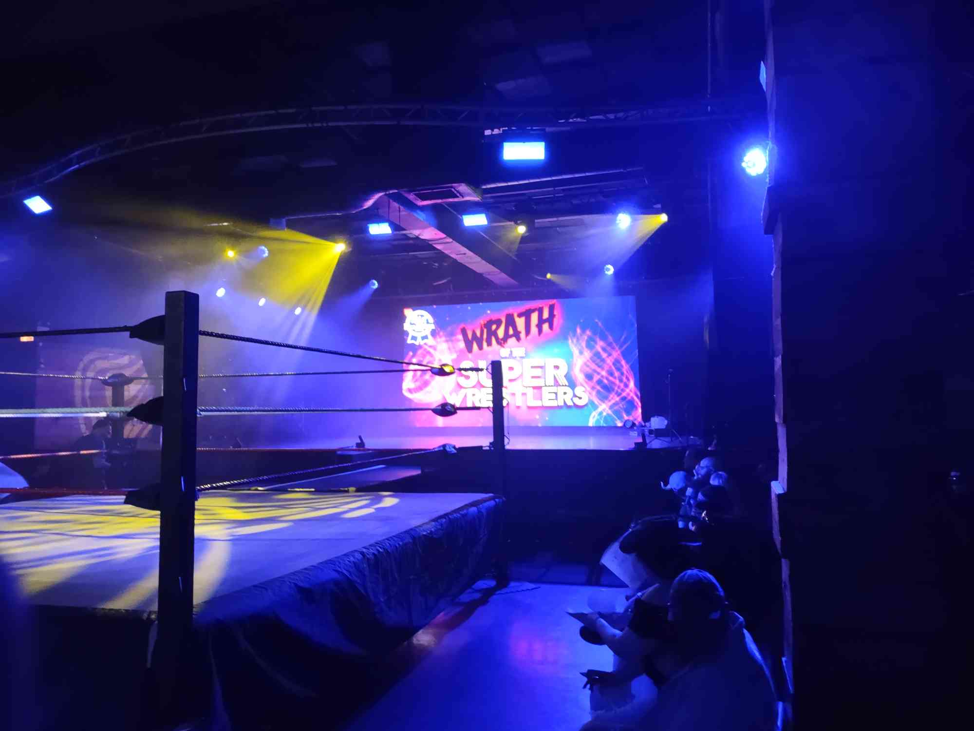a photo taken inside of a concert hall, a wrestling ring set up in the center of the room with a projector showing the text WRATH OF THE SUPER WRESTLERS